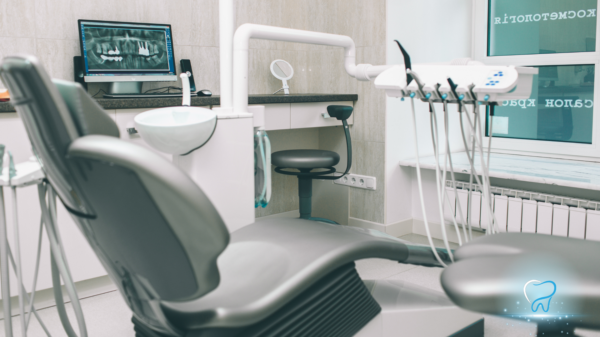 Modern dental clinic with gray chair, tools, sink, and monitor behind glass windows