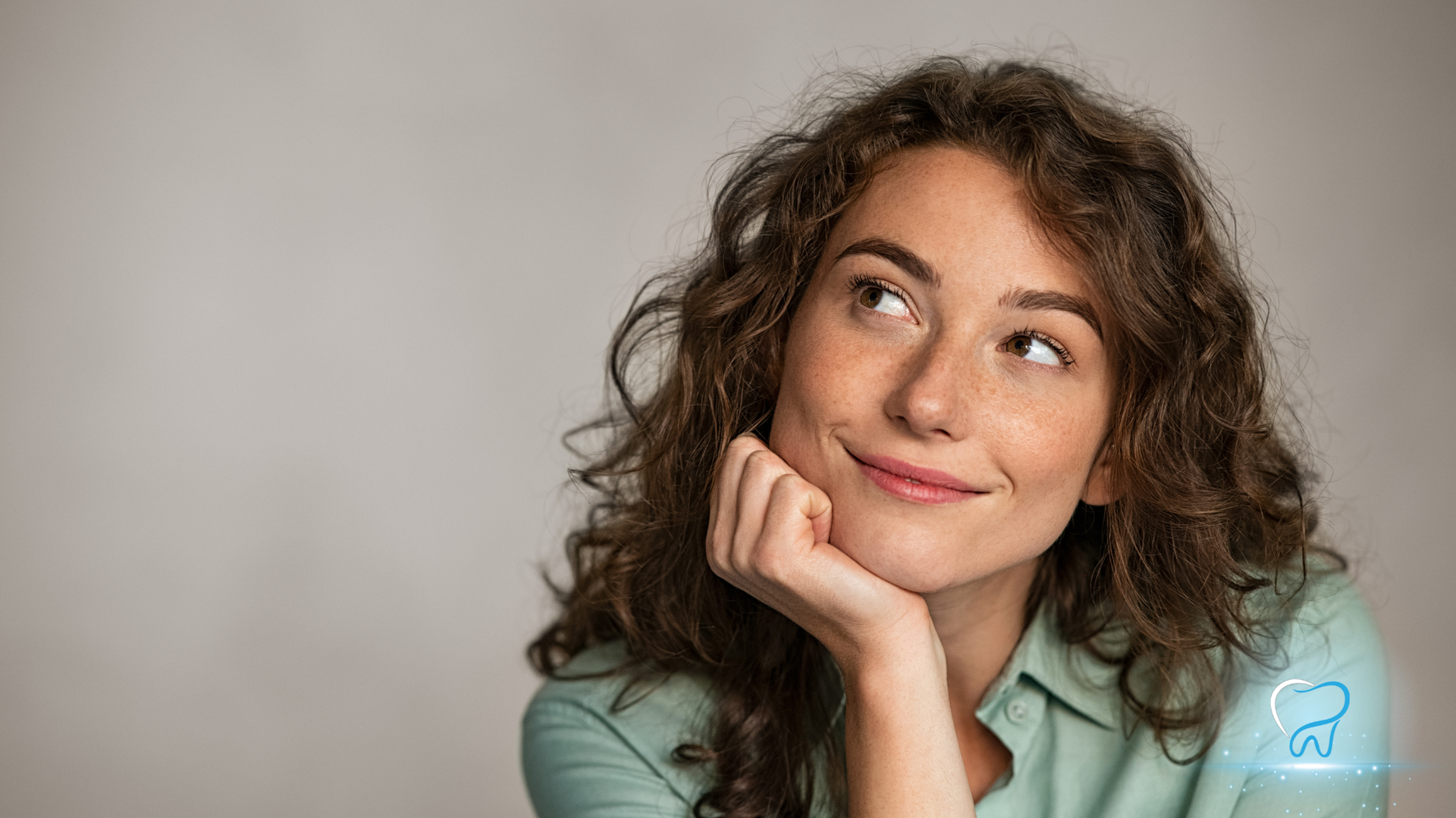 Person with curly hair resting chin on hand, smiling and looking upward against a plain background.