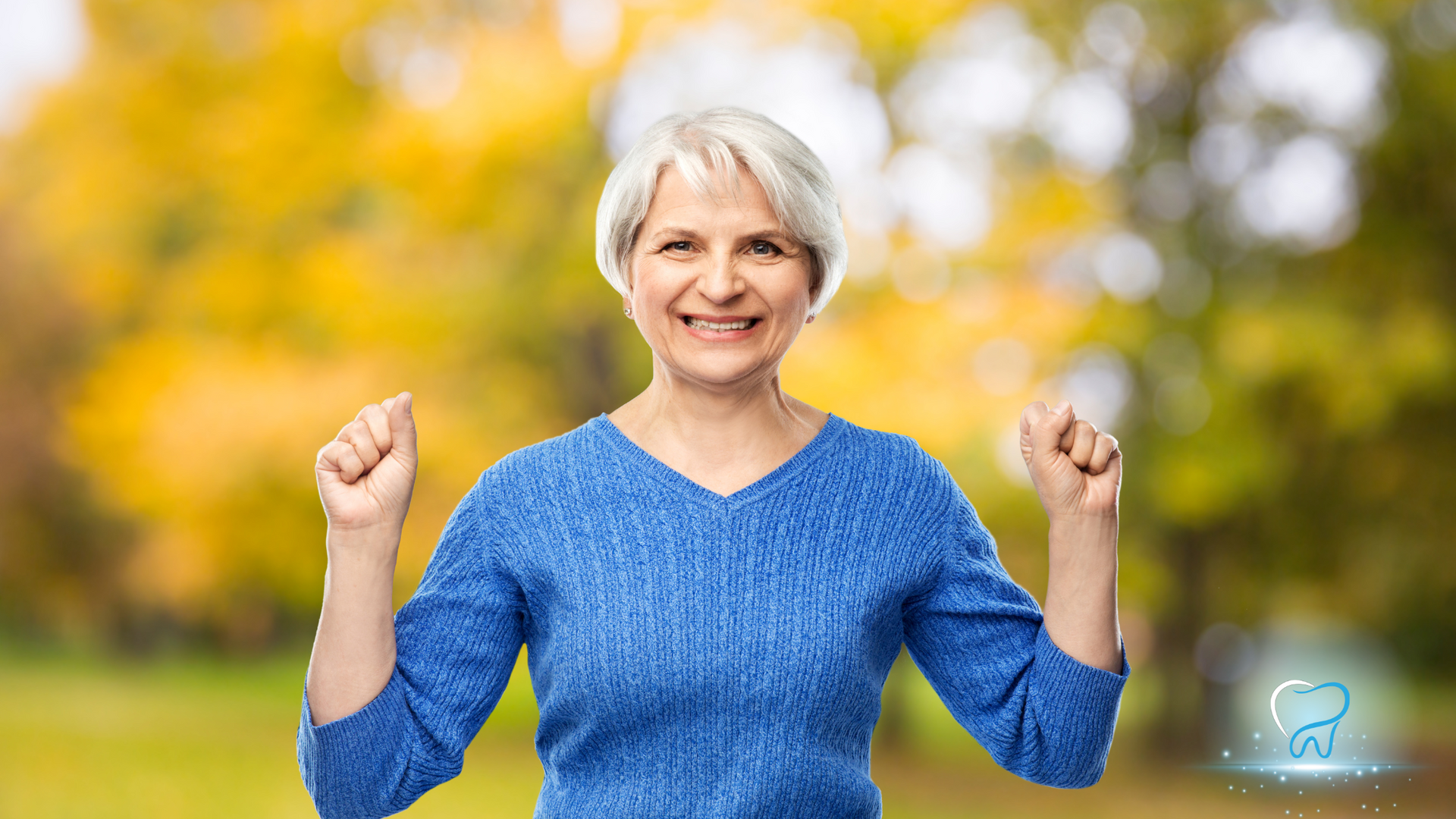 Smiling woman in a blue sweater raises both fists outdoors with blurred yellow trees behind her