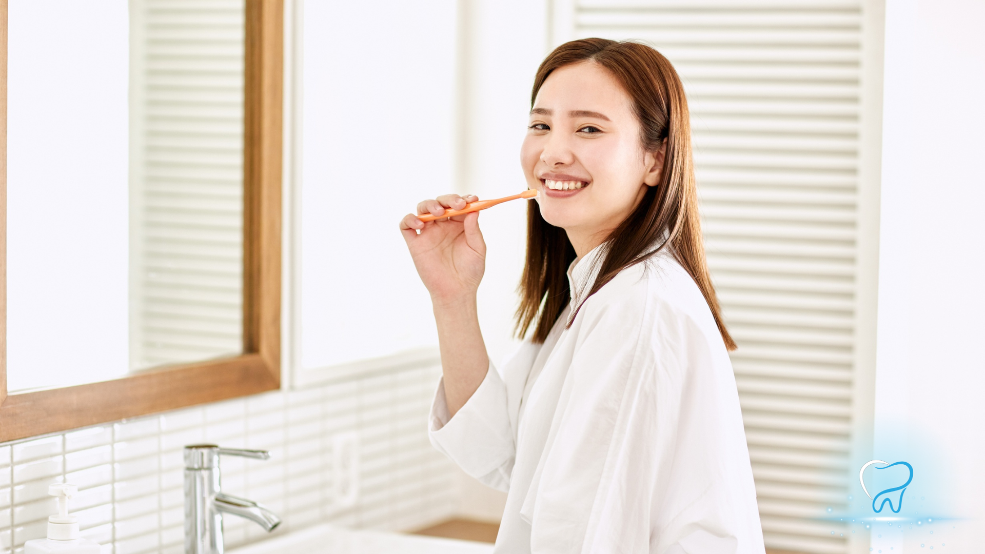 Person brushing teeth in a bright bathroom, smiling beside a sink and mirror.