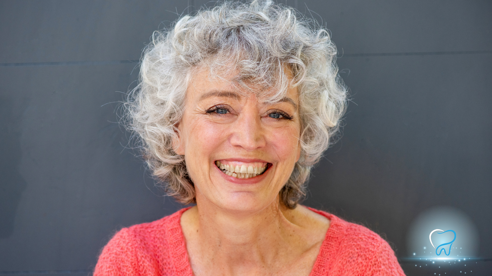 Smiling person with curly white hair in a coral top against a dark background