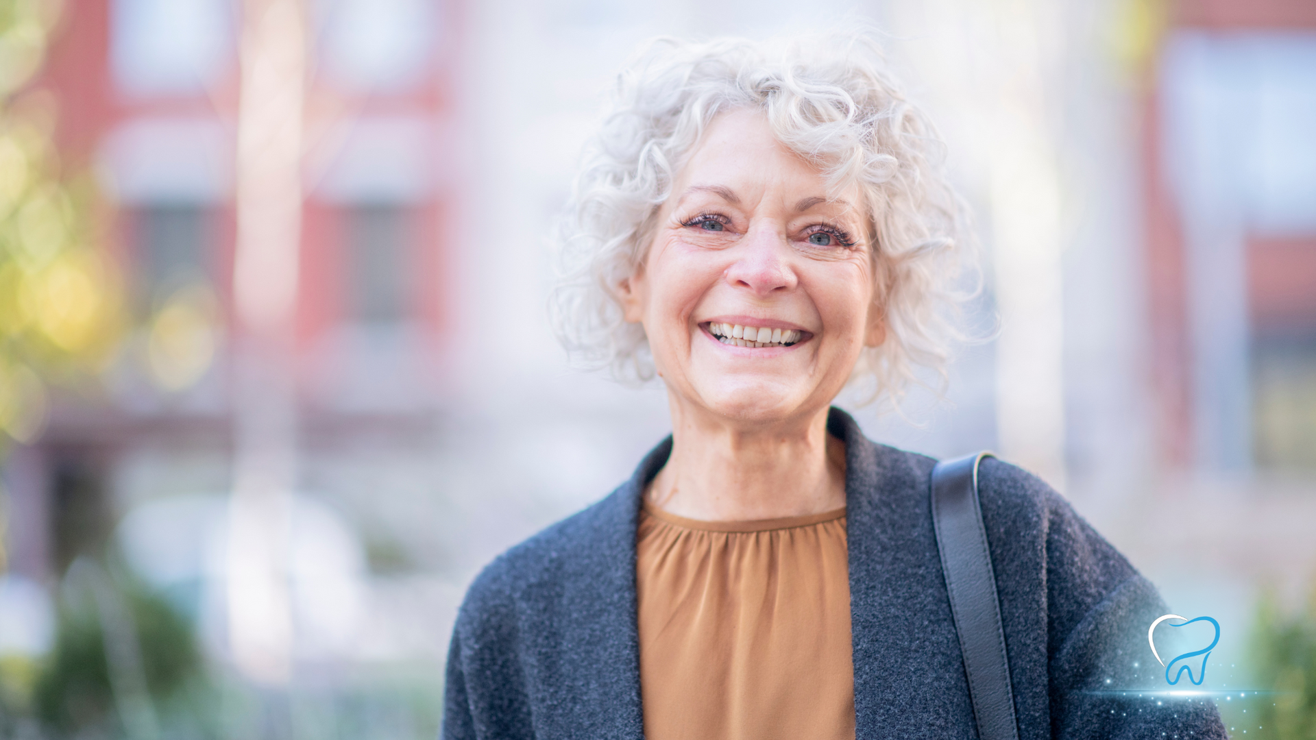 Smiling person with curly gray hair in a brown top and dark cardigan outdoors with blurred buildings behind.
