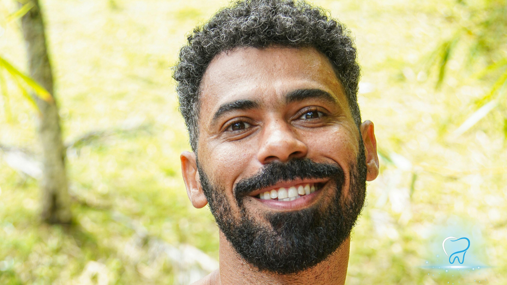 Smiling man with curly hair and beard outdoors against a bright green, blurred background
