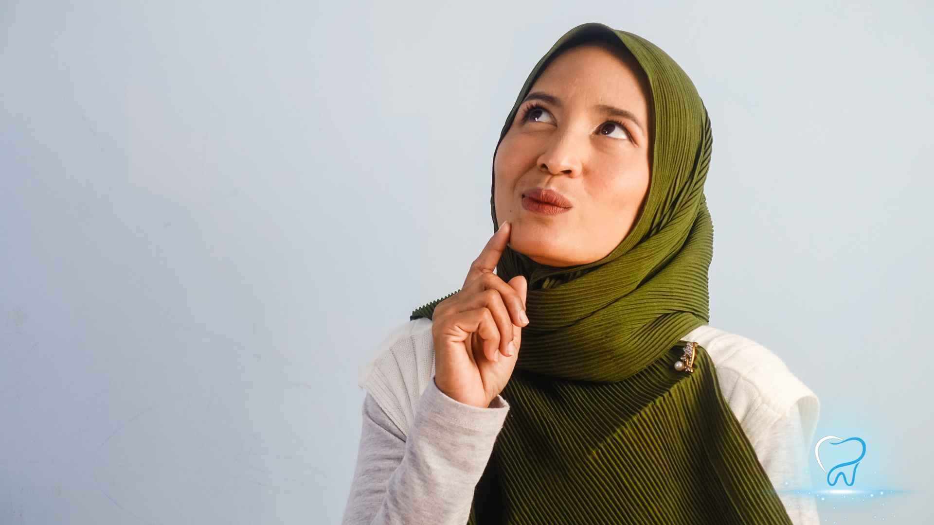 Woman in green hijab looking up thoughtfully, resting a finger on her chin against a plain light background