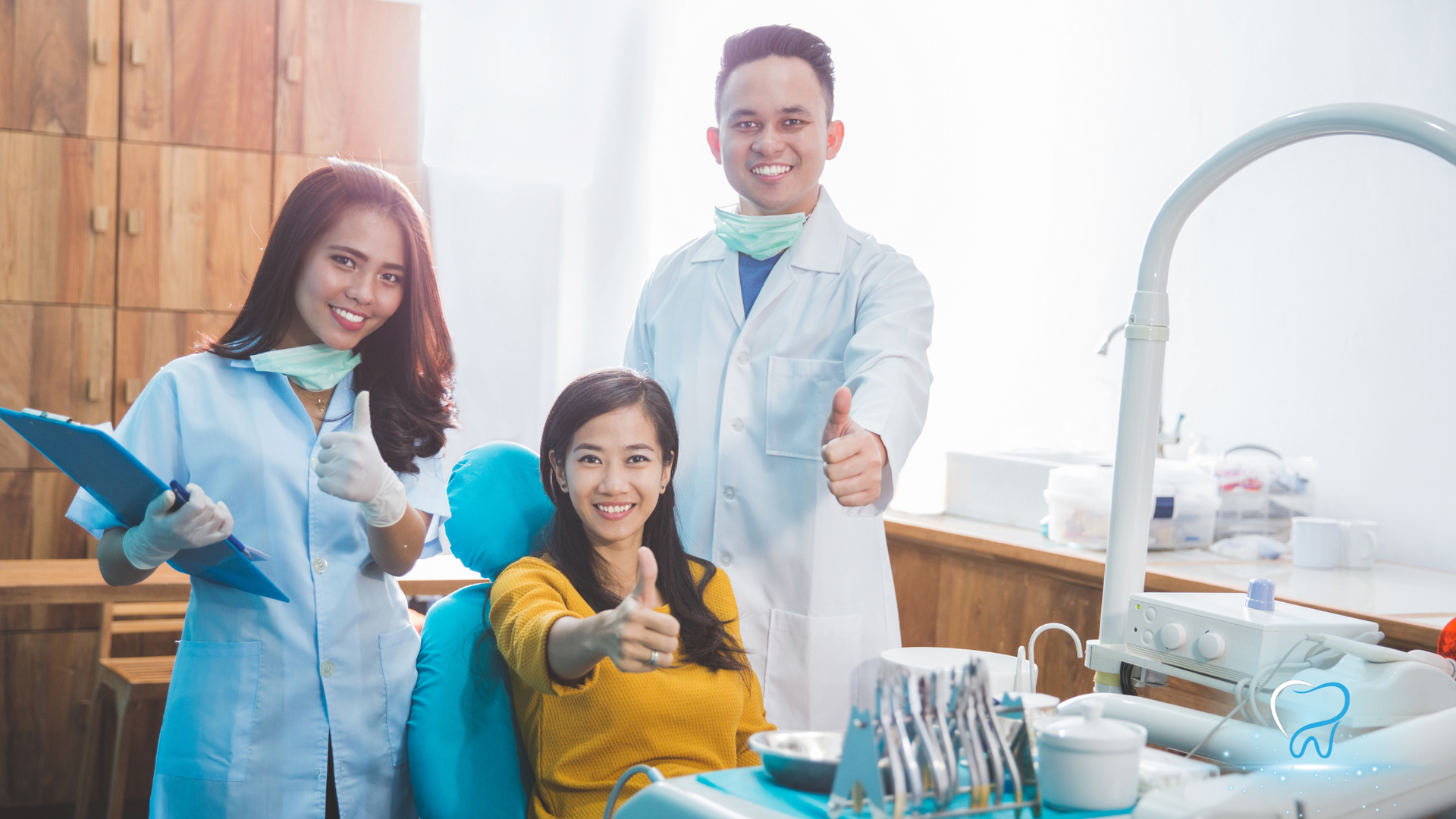 Three people in lab coats smiling in a bright dental clinic, giving thumbs up around dental tools.