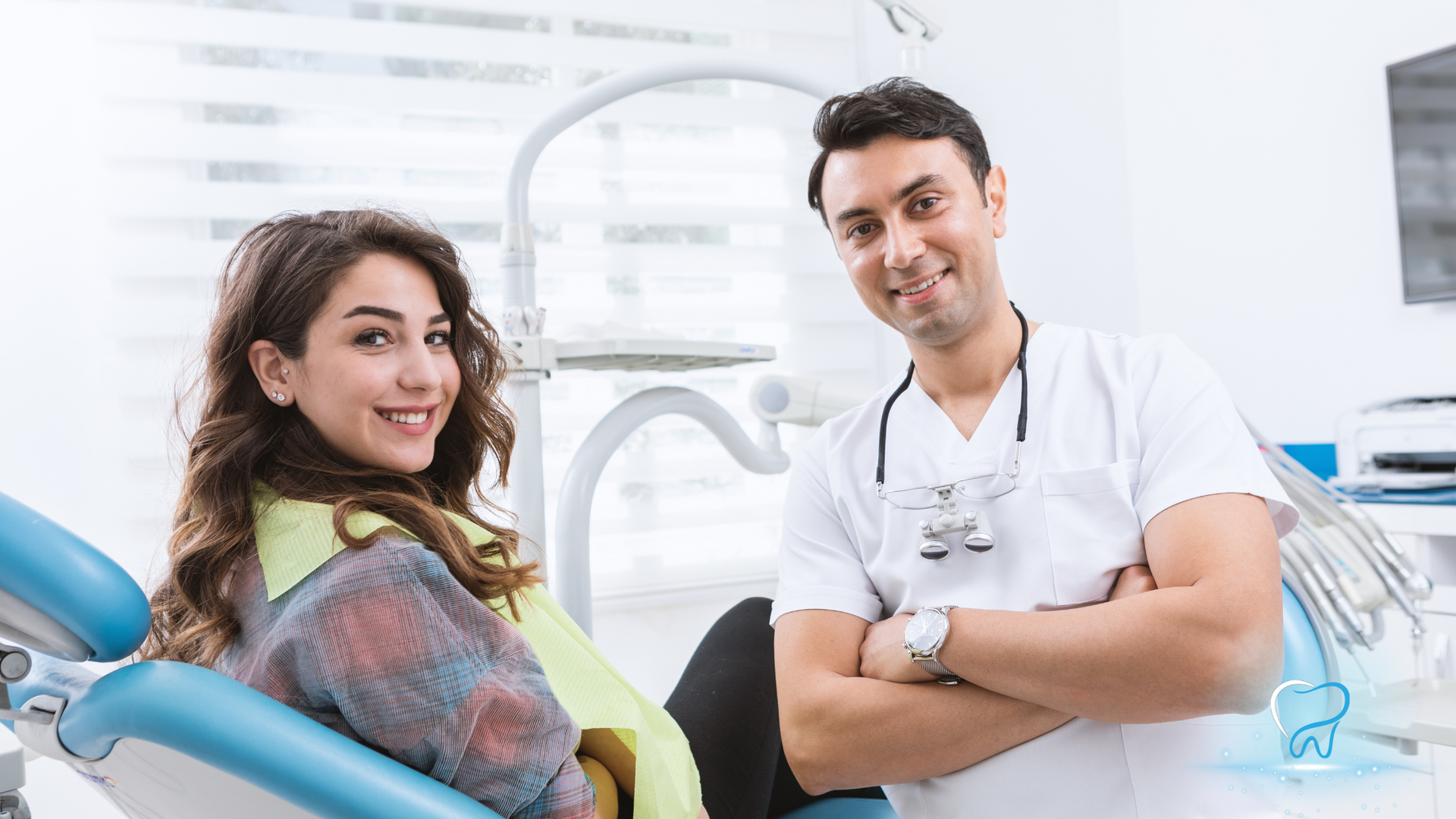 Dentist and patient smiling in a bright dental office, seated beside a blue dental chair.