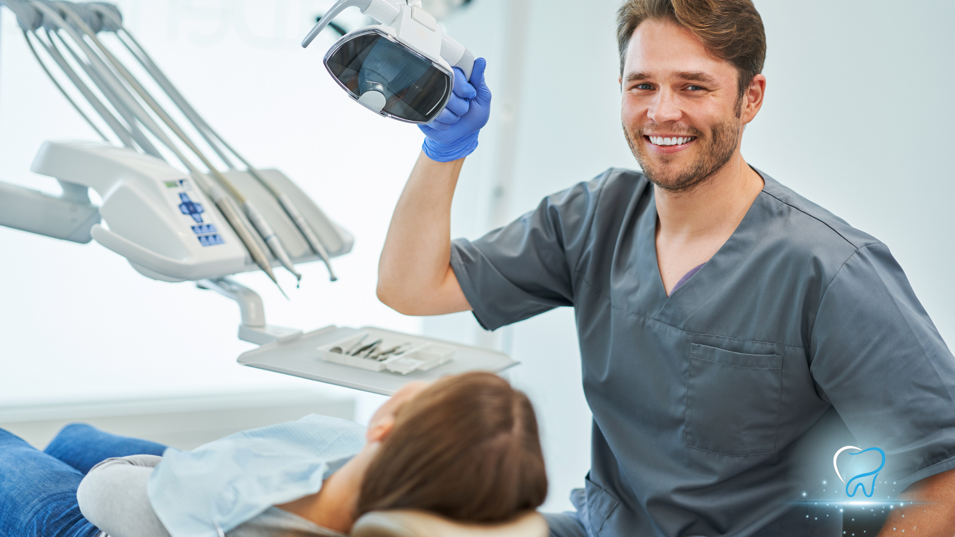 Dentist in gray scrubs holds a dental X-ray beside a patient in a bright clinic exam room