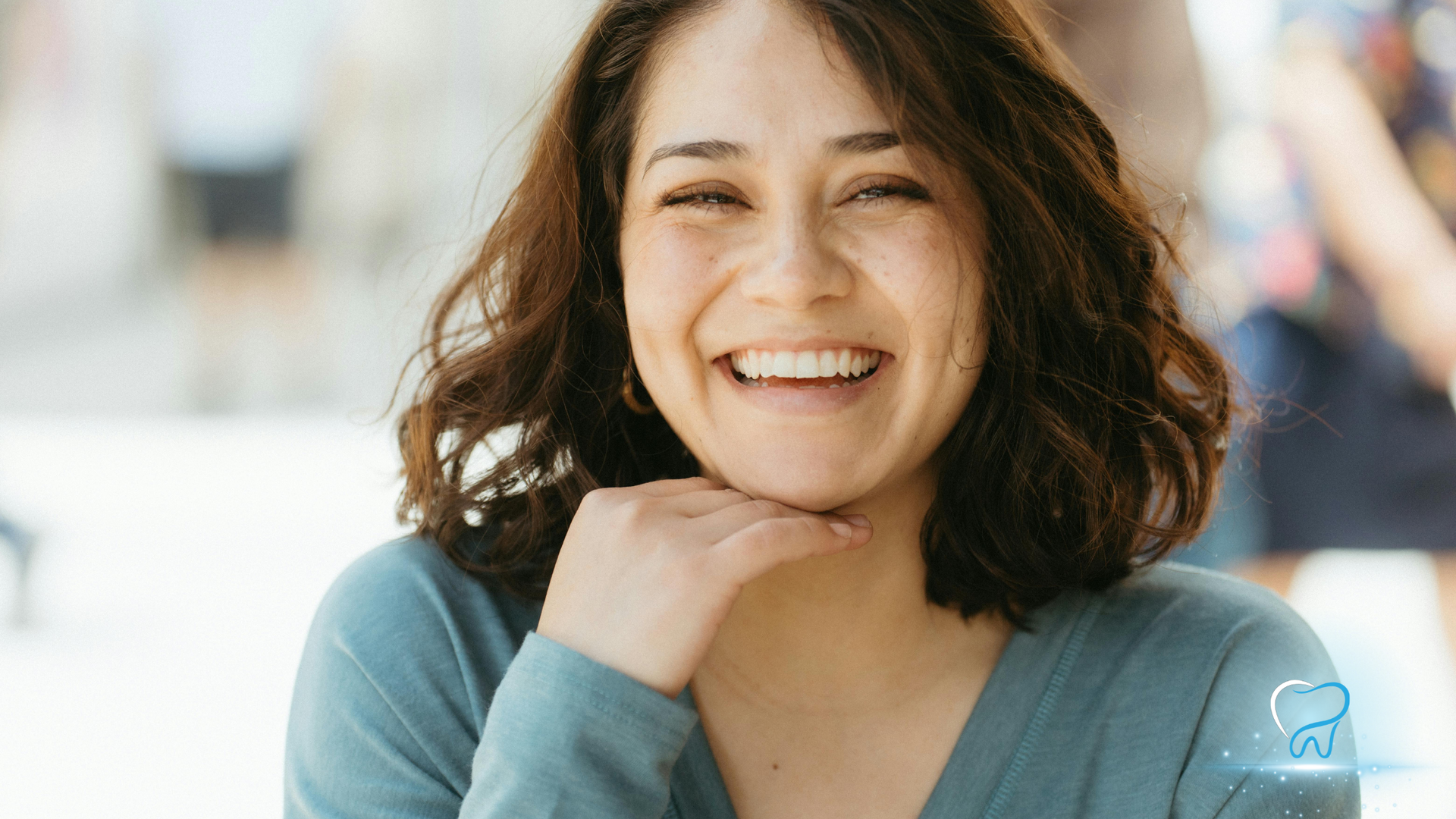 Smiling person with dark hair resting chin on hand, wearing a blue top against a bright blurred background