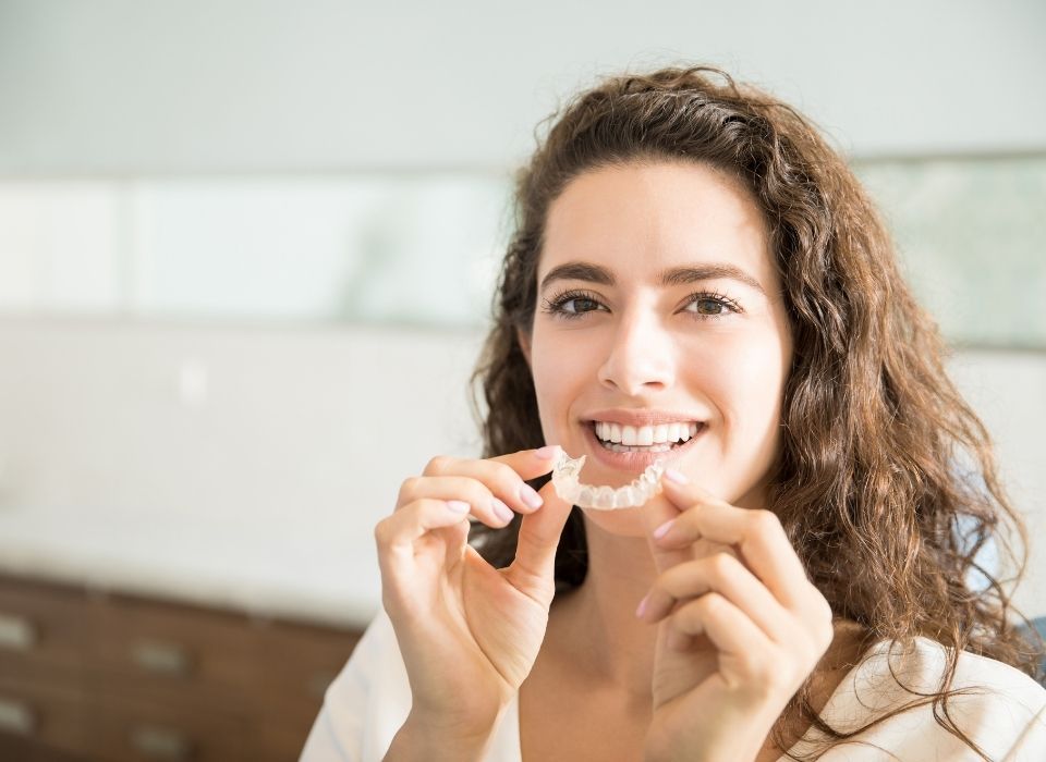 Woman holding clear aligners and smiling in a bright bathroom