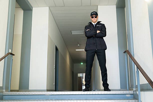 Security guard standing in a hallway, arms crossed, wearing uniform and sunglasses.
