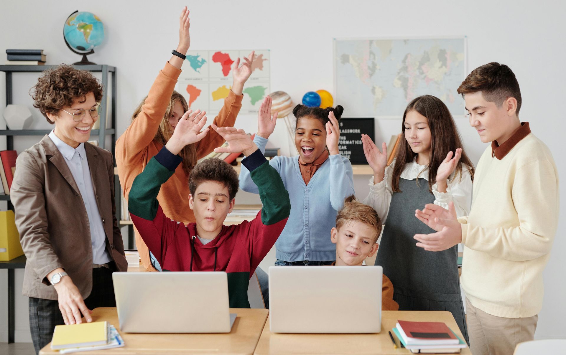 A group of children are standing around two laptops in a classroom.