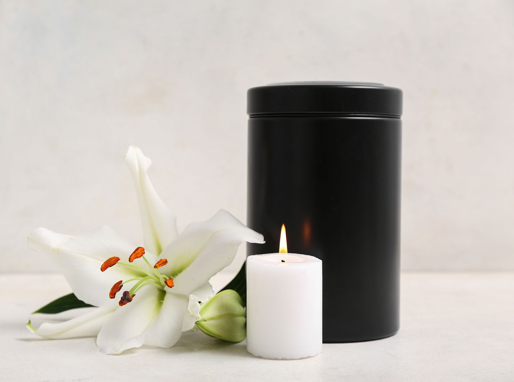 White lily and lit candle beside a black memorial urn on a white background