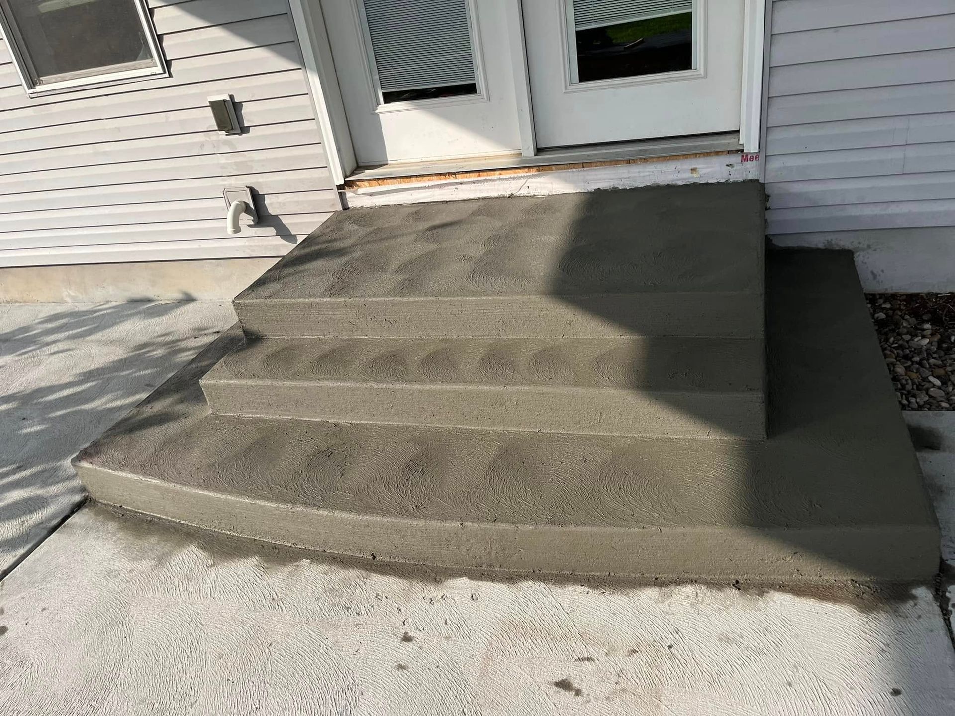 A set of newly poured, gray concrete steps leading to a white double door on a house with light-colored siding.