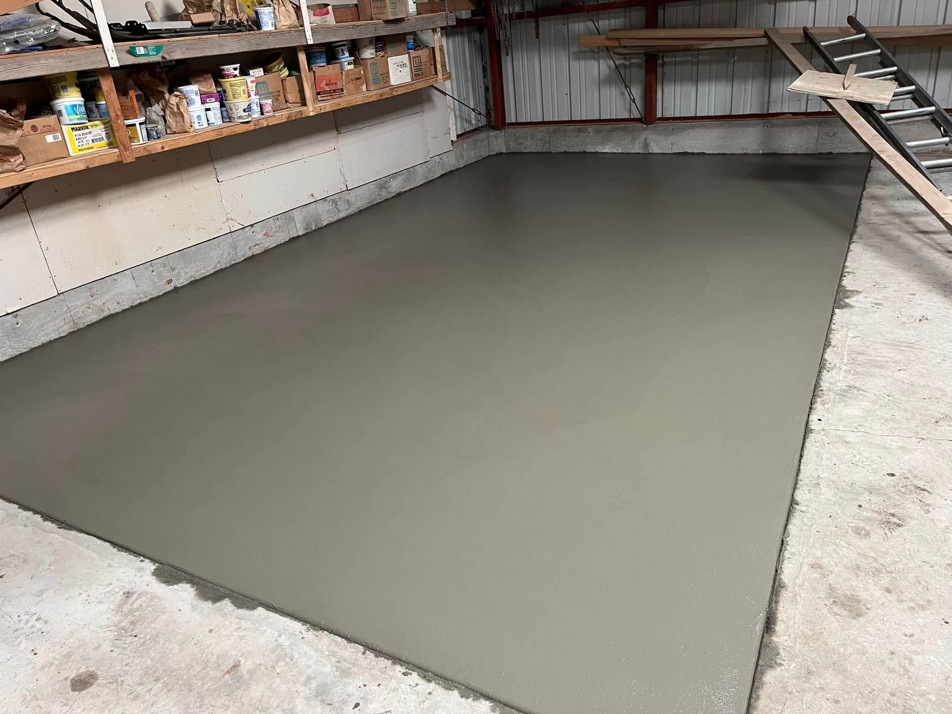 A newly poured, smooth gray concrete floor patch inside a workshop with shelving and a ladder in the background.