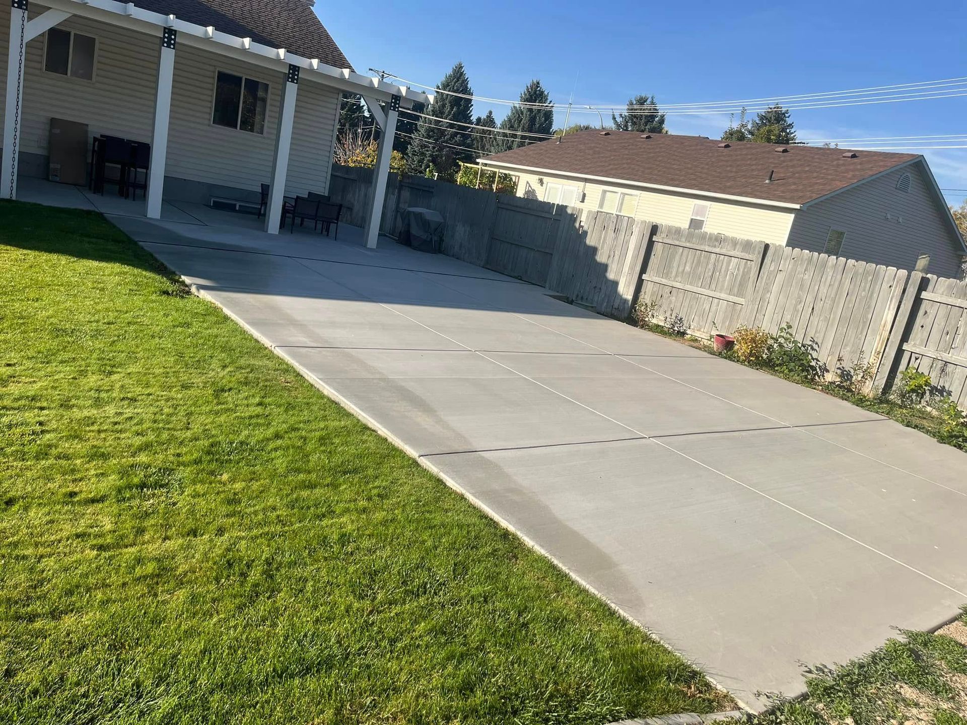 A large, rectangular concrete patio adjacent to a house under a covered porch, bordered by a lawn and a concrete fence.