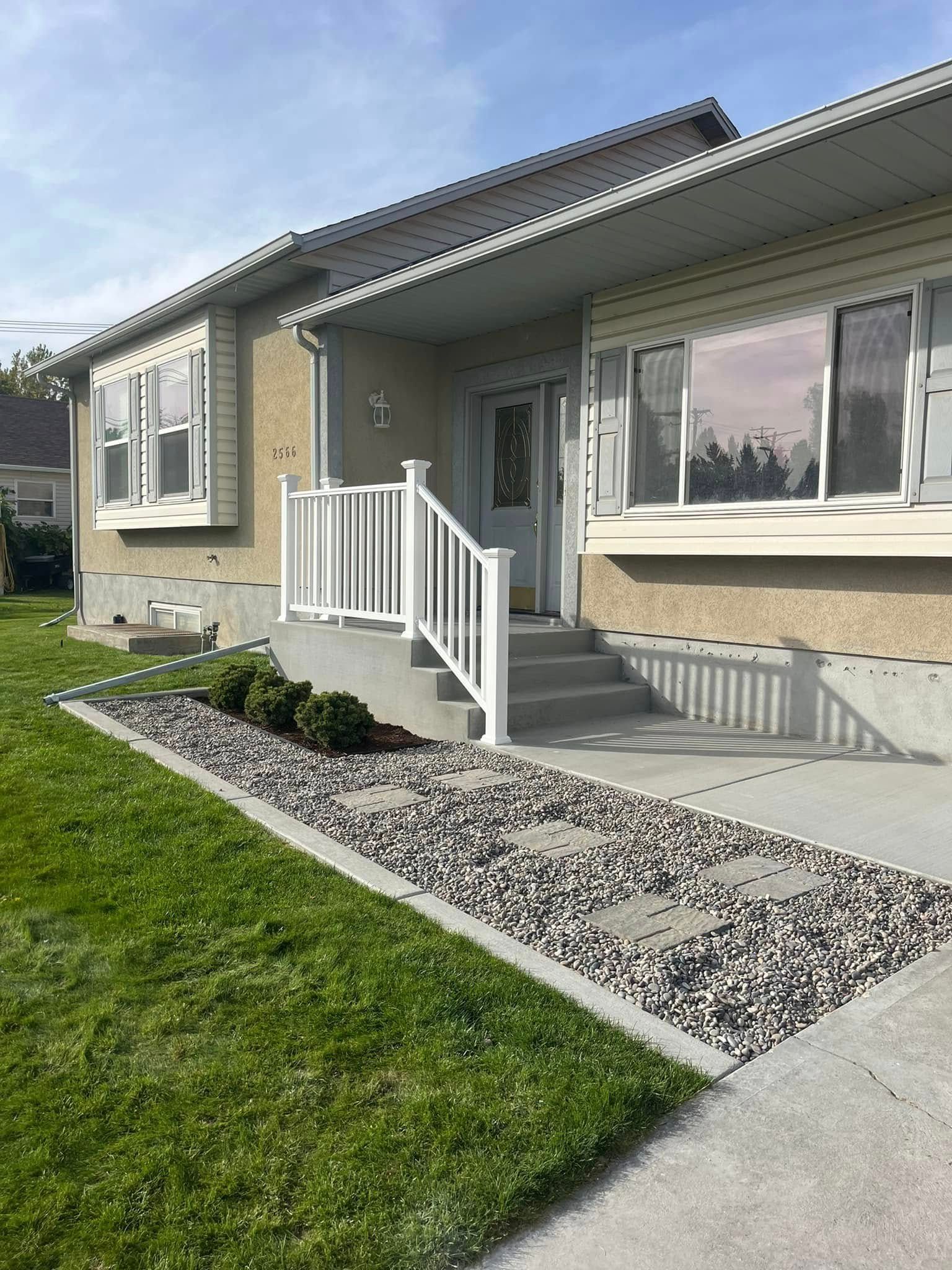 A suburban house with a tan stucco exterior, white railing on the front stairs, and a gravel pathway beside a green lawn.