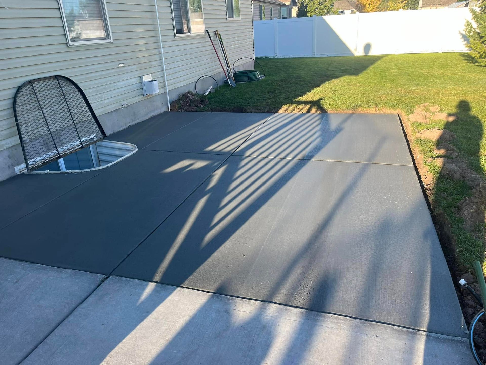 A newly poured gray concrete patio sits against the side of a house next to a basement window well with a metal cover.
