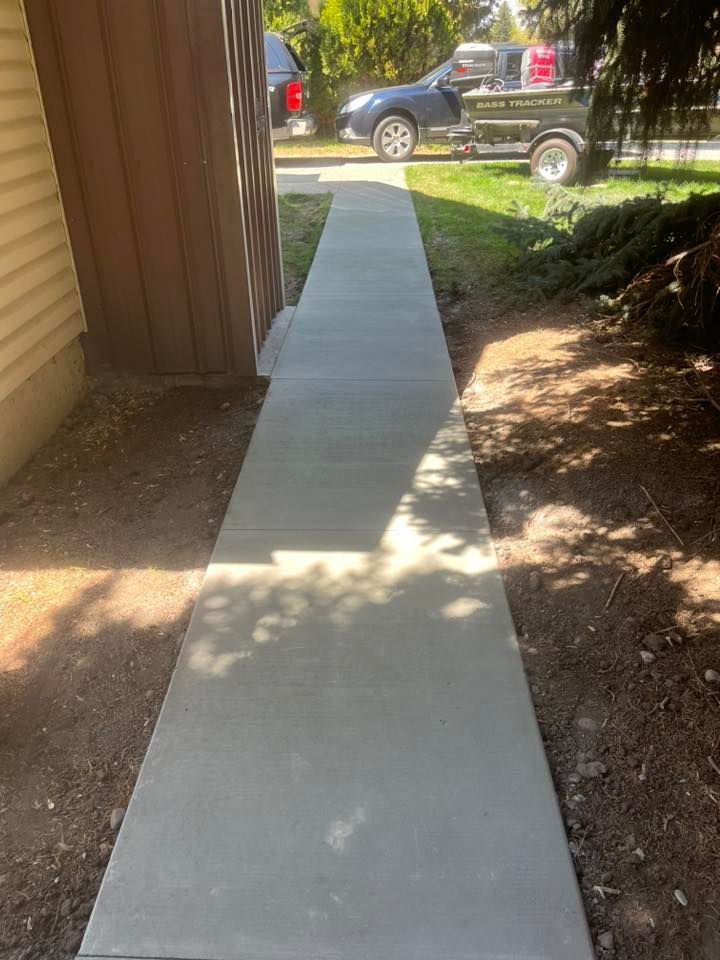 A newly poured concrete walkway stretching between a house exterior and a patch of ground toward a driveway.