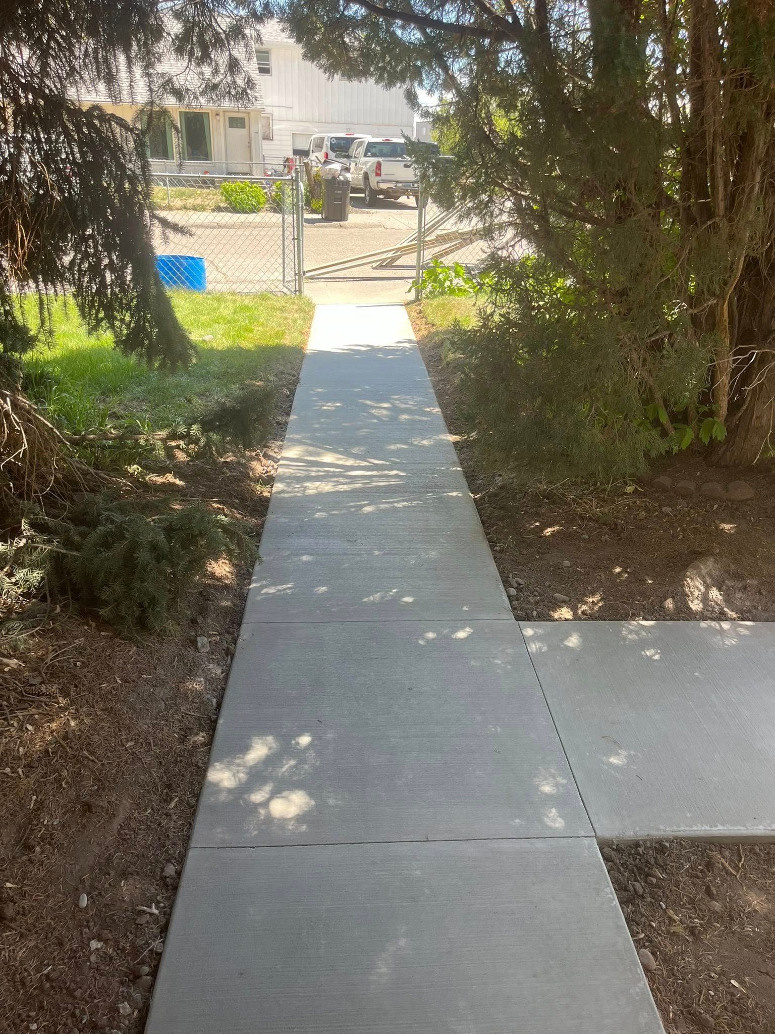 A concrete sidewalk pathway leads through a yard toward a gated driveway with a parked vehicle in the background.