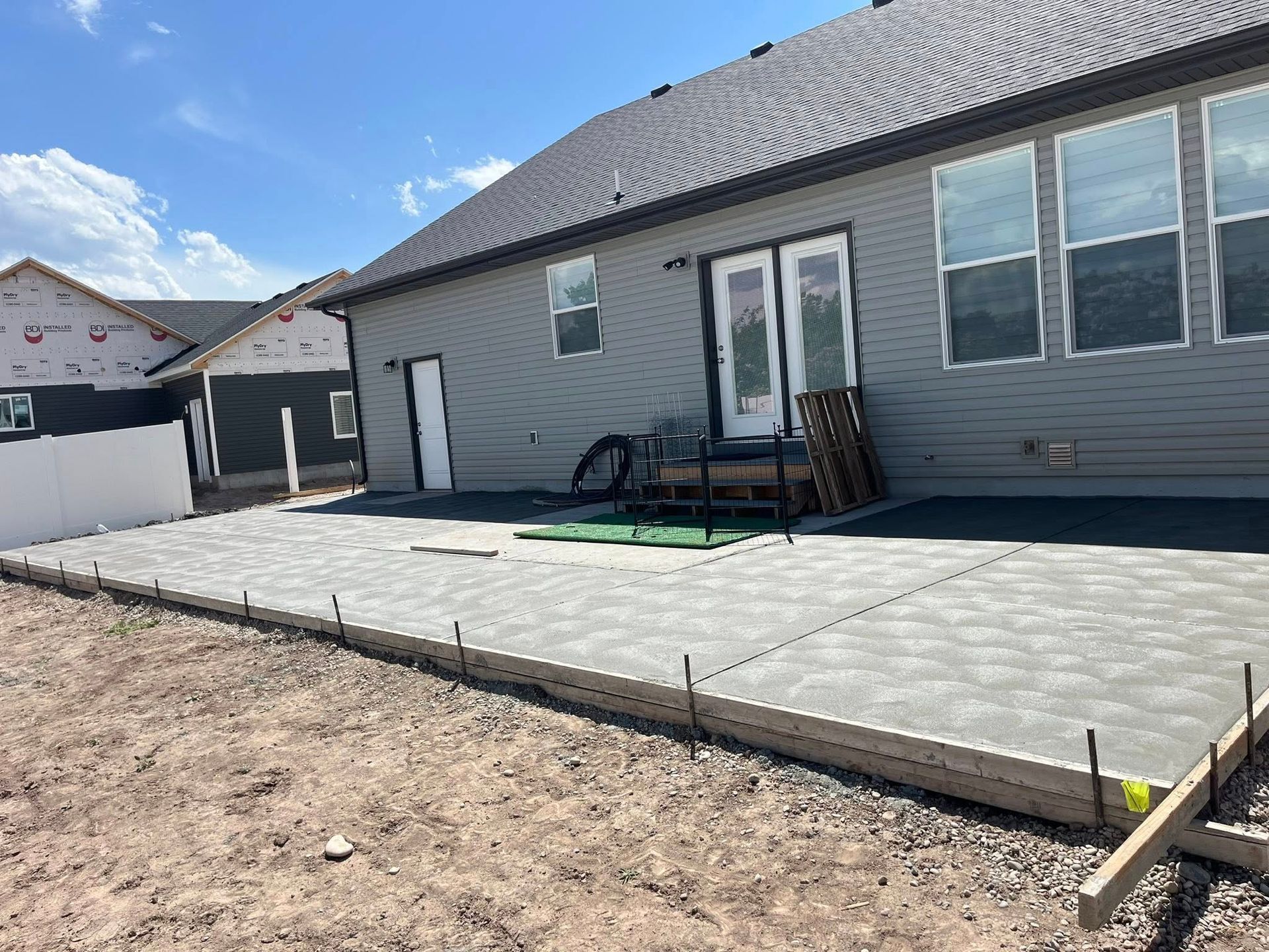 A newly poured concrete patio slab outside the back of a grey, two-story residential house under a bright blue sky.