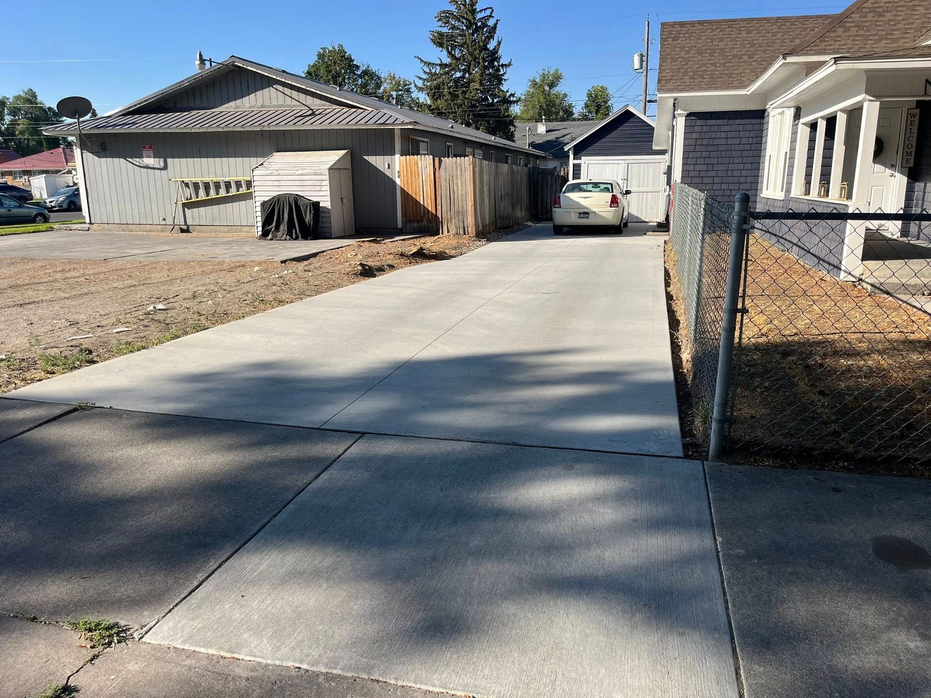 A concrete driveway leads toward a house and a light-colored car parked behind a wooden fence under a clear blue sky.