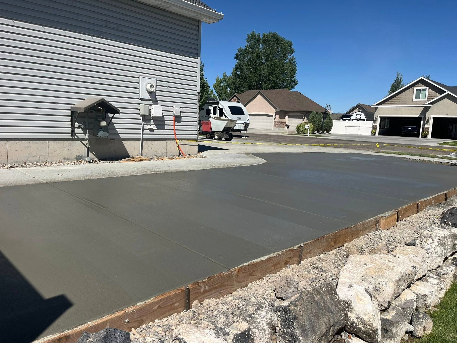 Newly poured concrete driveway beside a house with a rock retaining wall and a parked camper in the background.