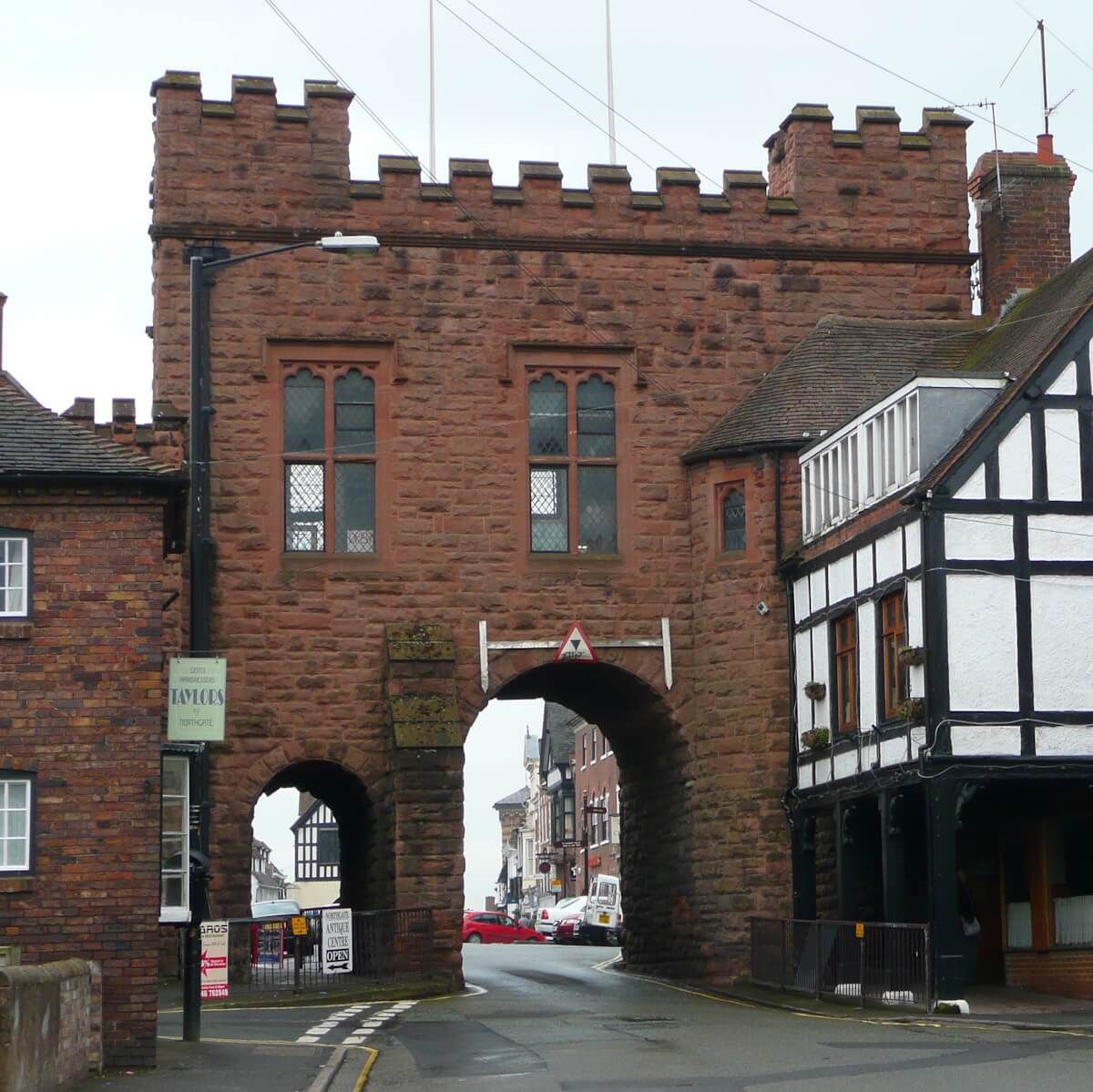 Red sandstone archway with crenellations; road leads through it. Tudor-style building on the right.
