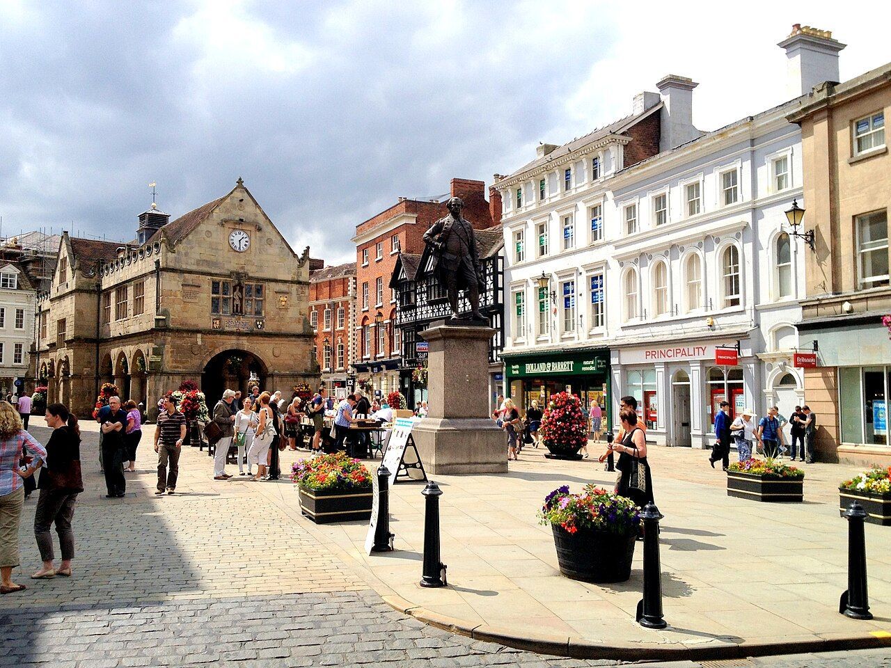 Shrewsbury street scene in front of historic buildings. People walking around a monument and shops.
