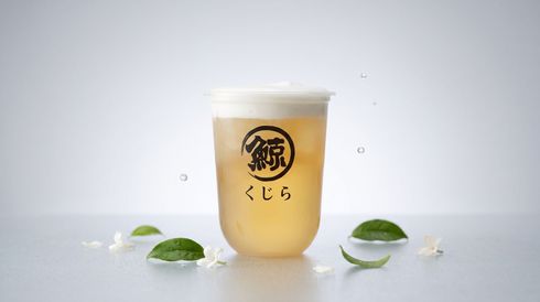 Transparent cup of beige milk tea with Japanese logo, surrounded by green mochi on a white surface