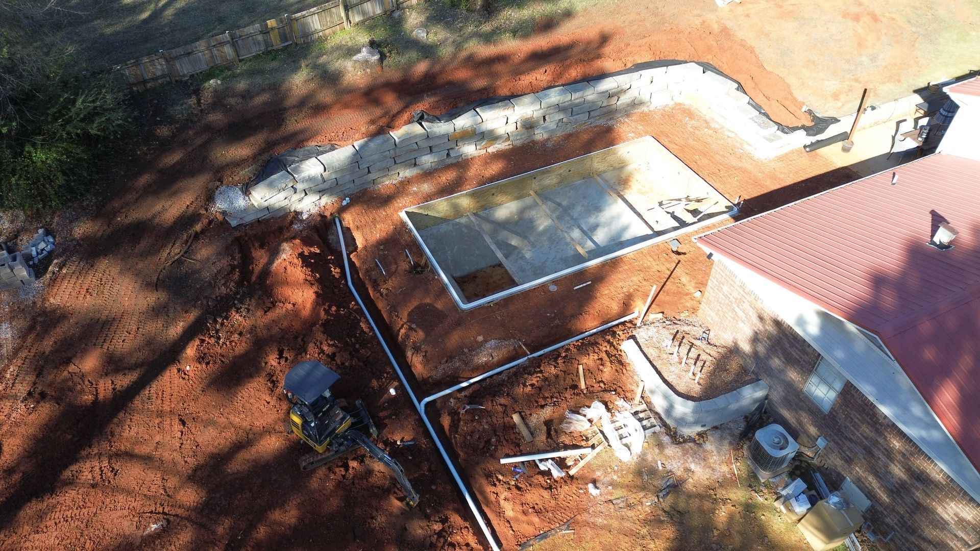 Aerial view of a residential backyard with a partially excavated swimming pool, red soil, and construction machinery.