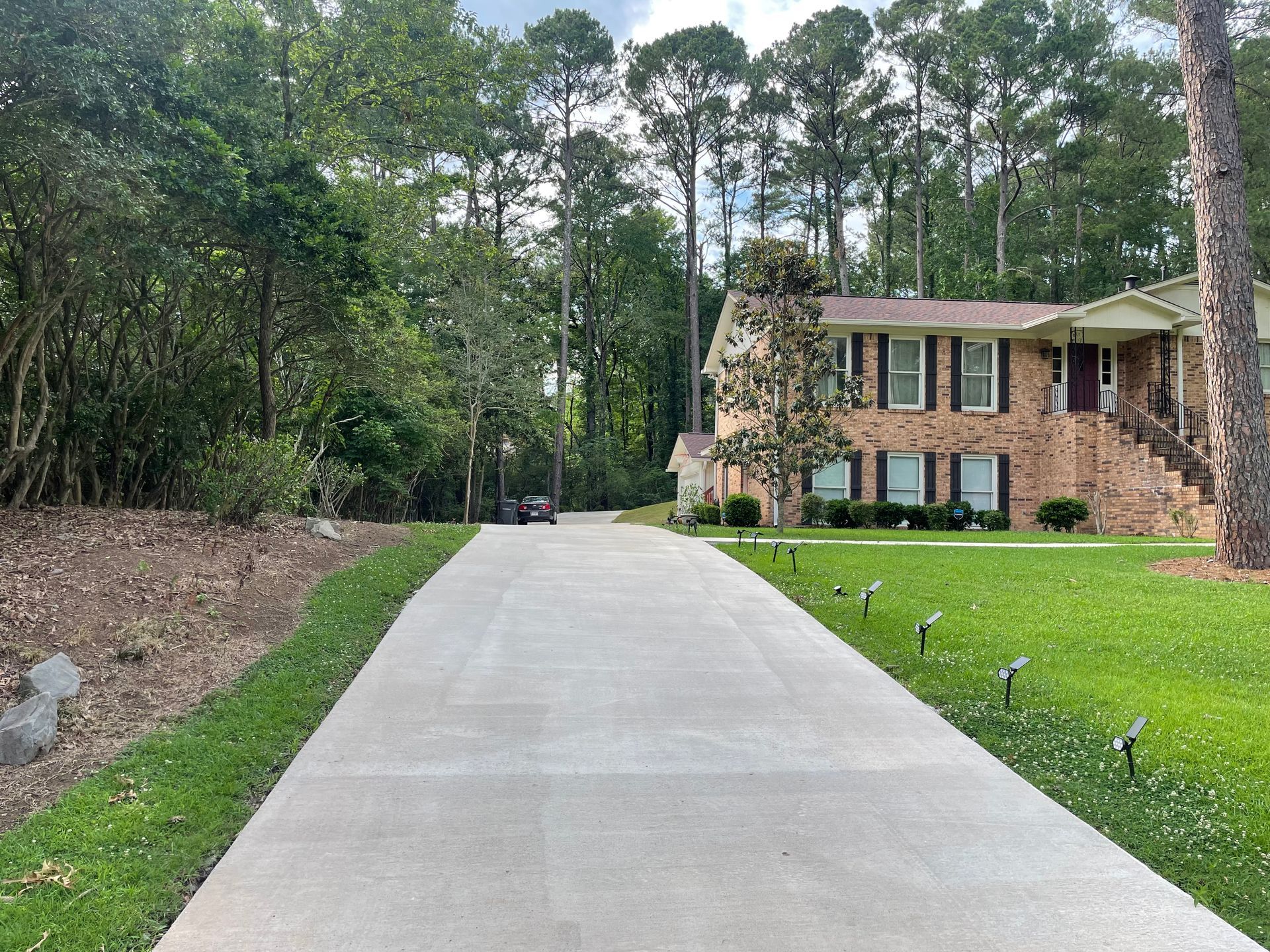A concrete driveway leads to a brick, two-story house surrounded by tall pine trees and a grassy lawn with solar lights.