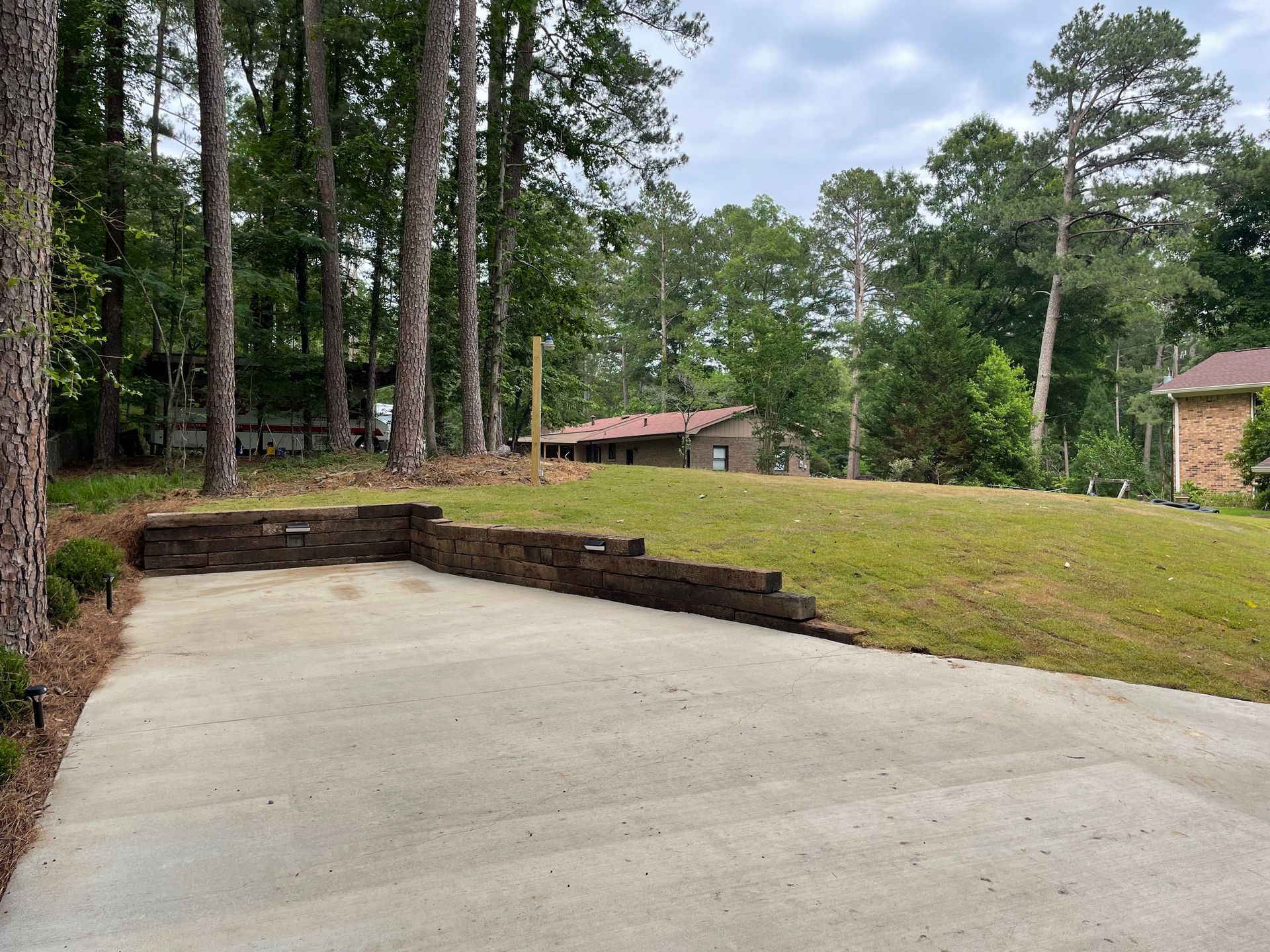 A concrete driveway area with a stone retaining wall bordering a grassy yard and surrounding trees under a cloudy sky.