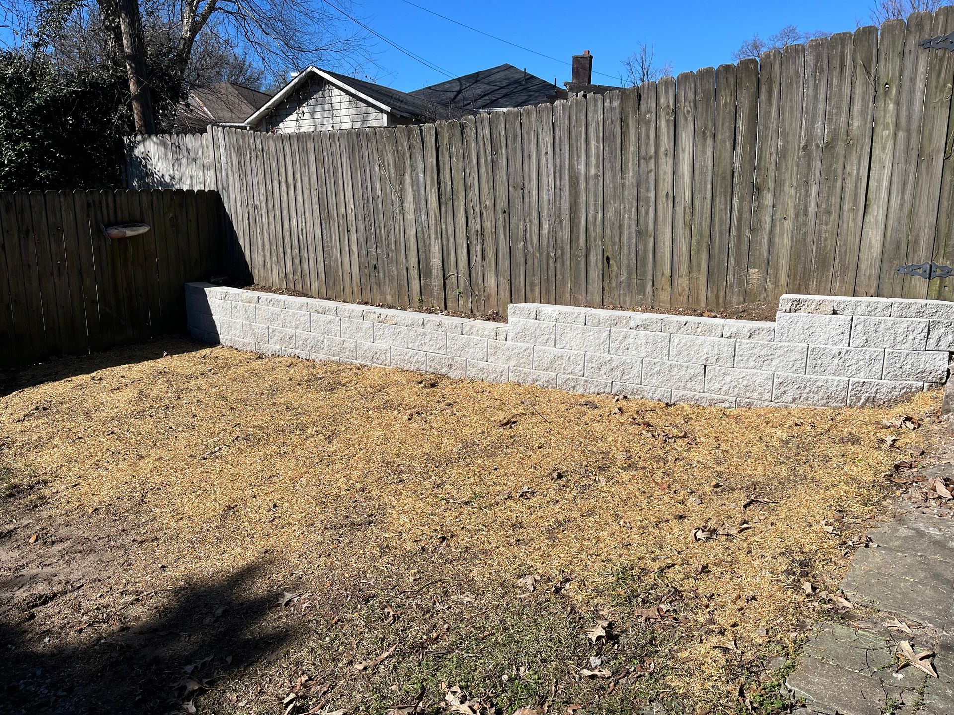 A backyard area with a new grey stone retaining wall built against a wooden fence, surrounded by mulch and dirt.
