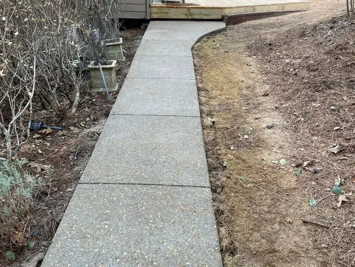 A concrete walkway leads toward a wooden deck, flanked by shrubs on the left and a dirt slope on the right.