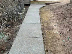 A concrete sidewalk leads to a wooden deck, flanked by shrubs on the left and a dirt slope on the right.