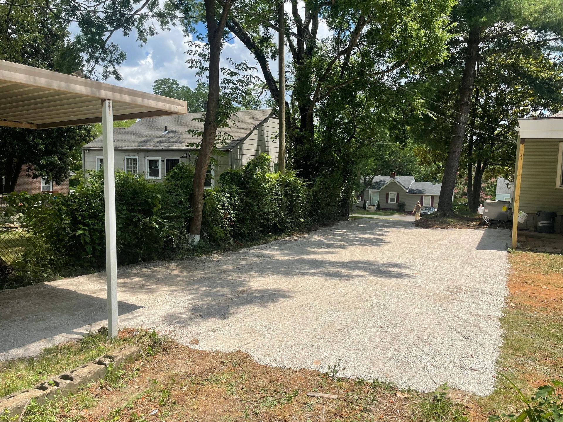 A gravel driveway leads toward a house, partially shaded by a carport structure in the foreground and surrounding trees.