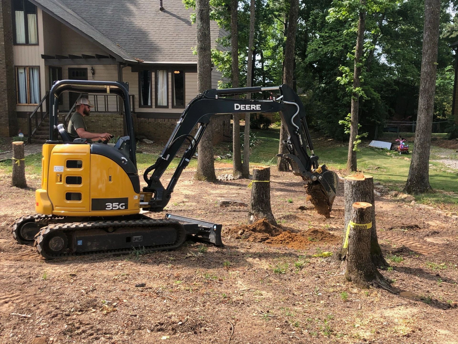 A yellow Deere mini excavator with its bucket in the ground among several tree stumps in a residential yard.