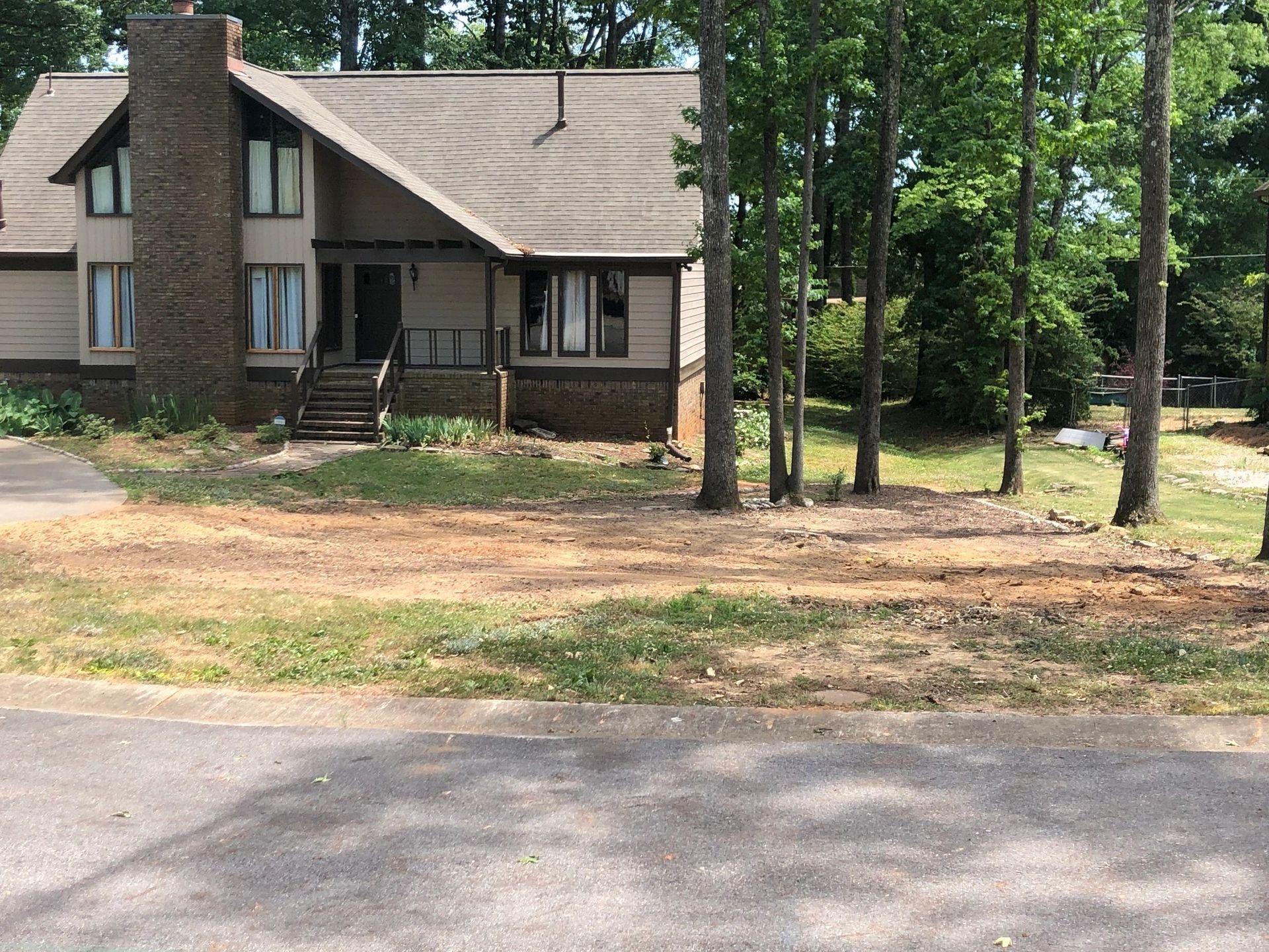A split-level house with brown siding and a stone chimney sits behind a cleared, dirt-covered yard and trees.