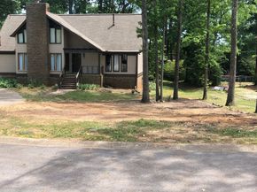 A two-story beige house with a brick chimney stands behind a large, cleared dirt yard with surrounding trees.