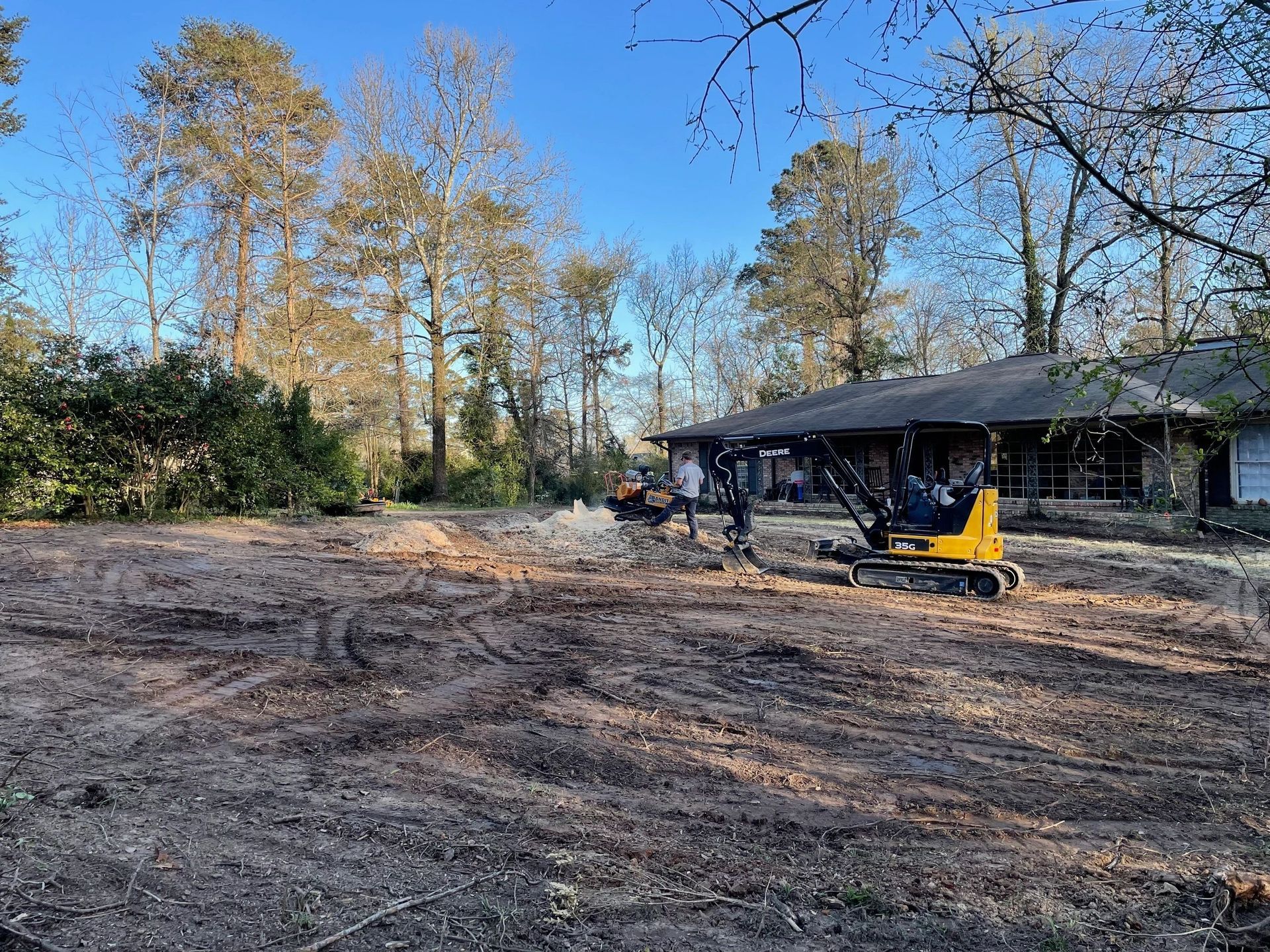 A yellow excavator and workers on a cleared lot with tire tracks in the dirt, near a house and trees under a clear sky.