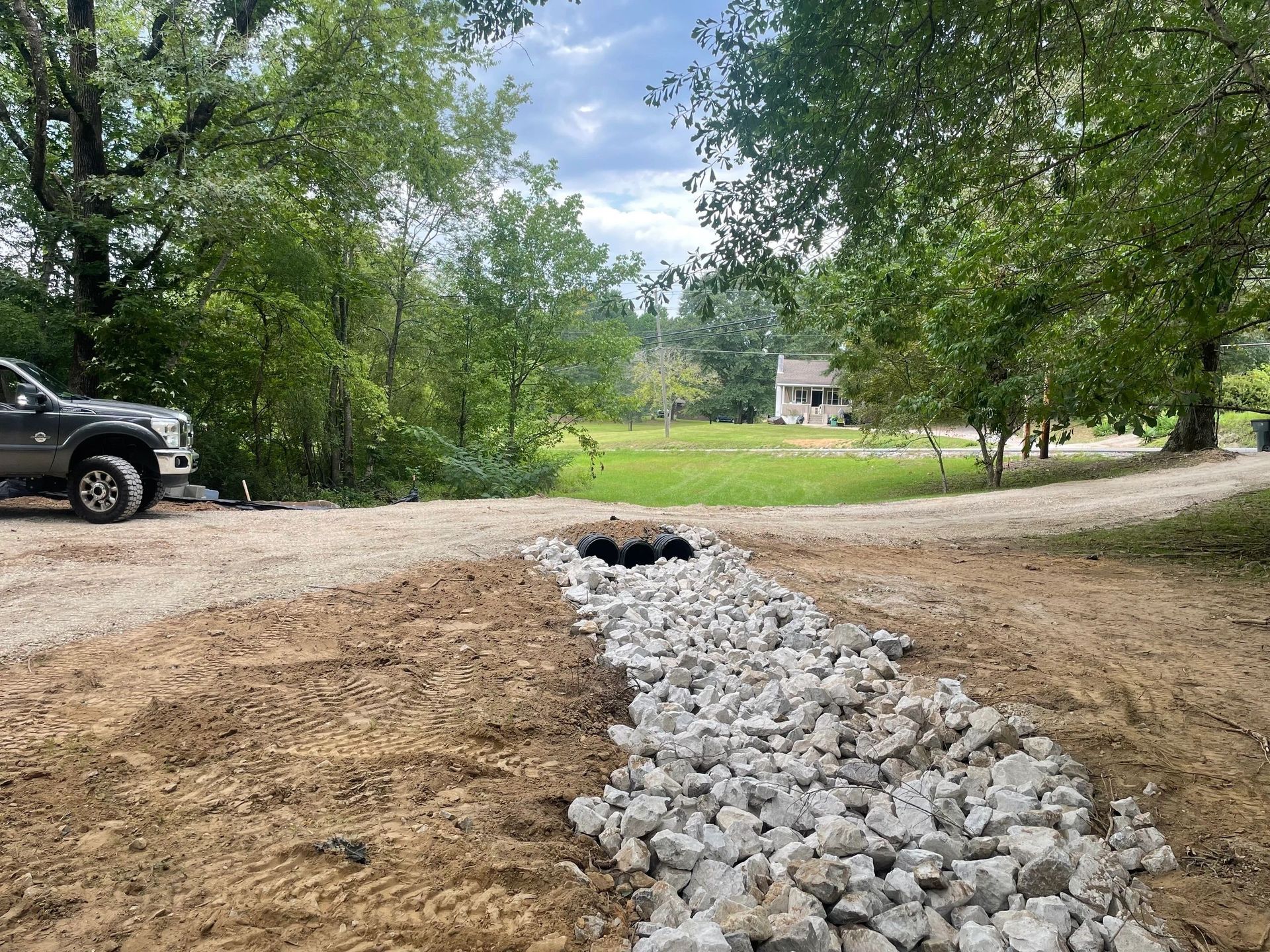 A gravel-lined drainage trench leading to culvert pipes under a gravel driveway, with trees and a house in the background.