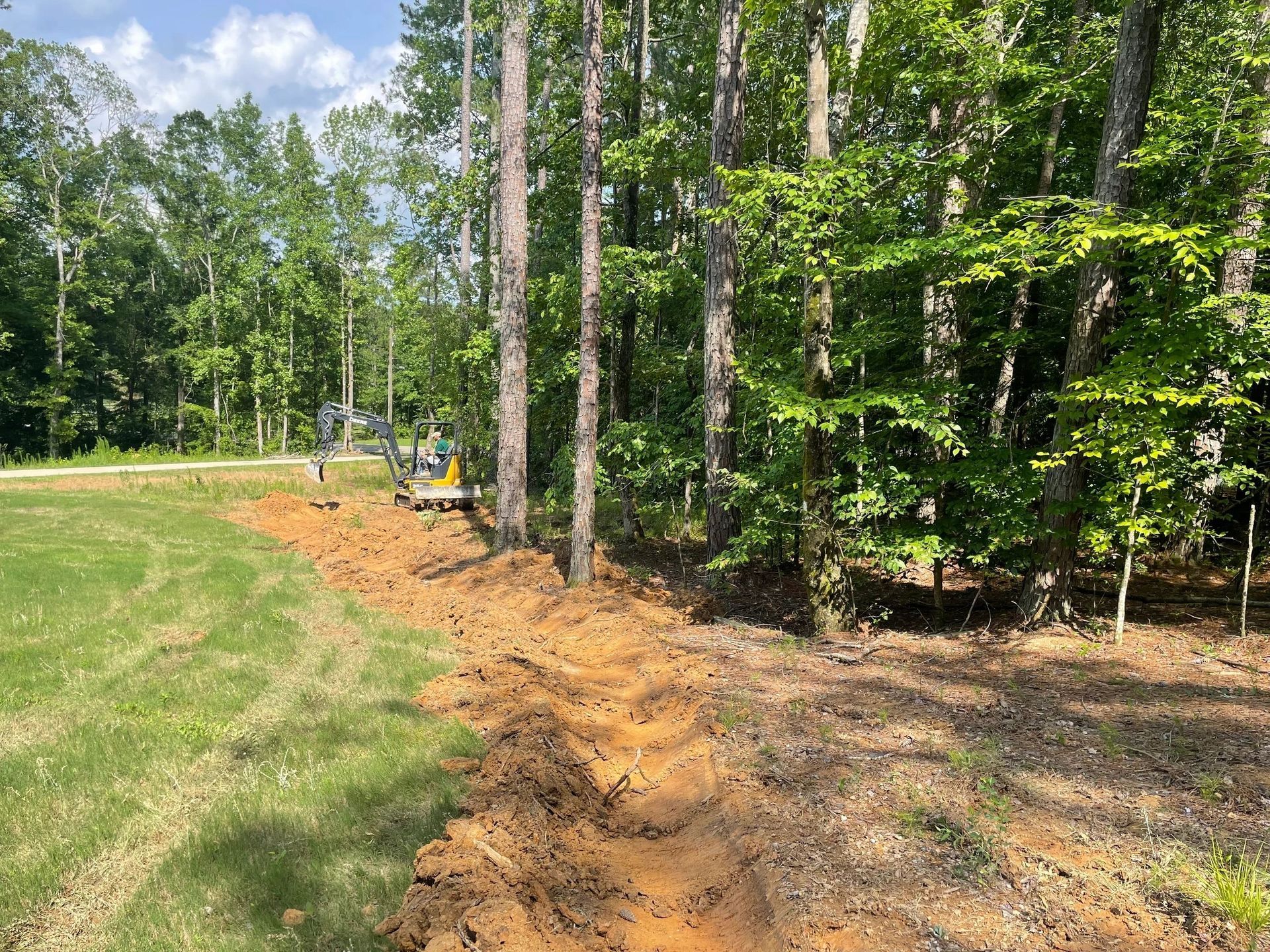 A construction excavator digs a long, narrow trench in the earth along the edge of a grassy lawn and a forest.
