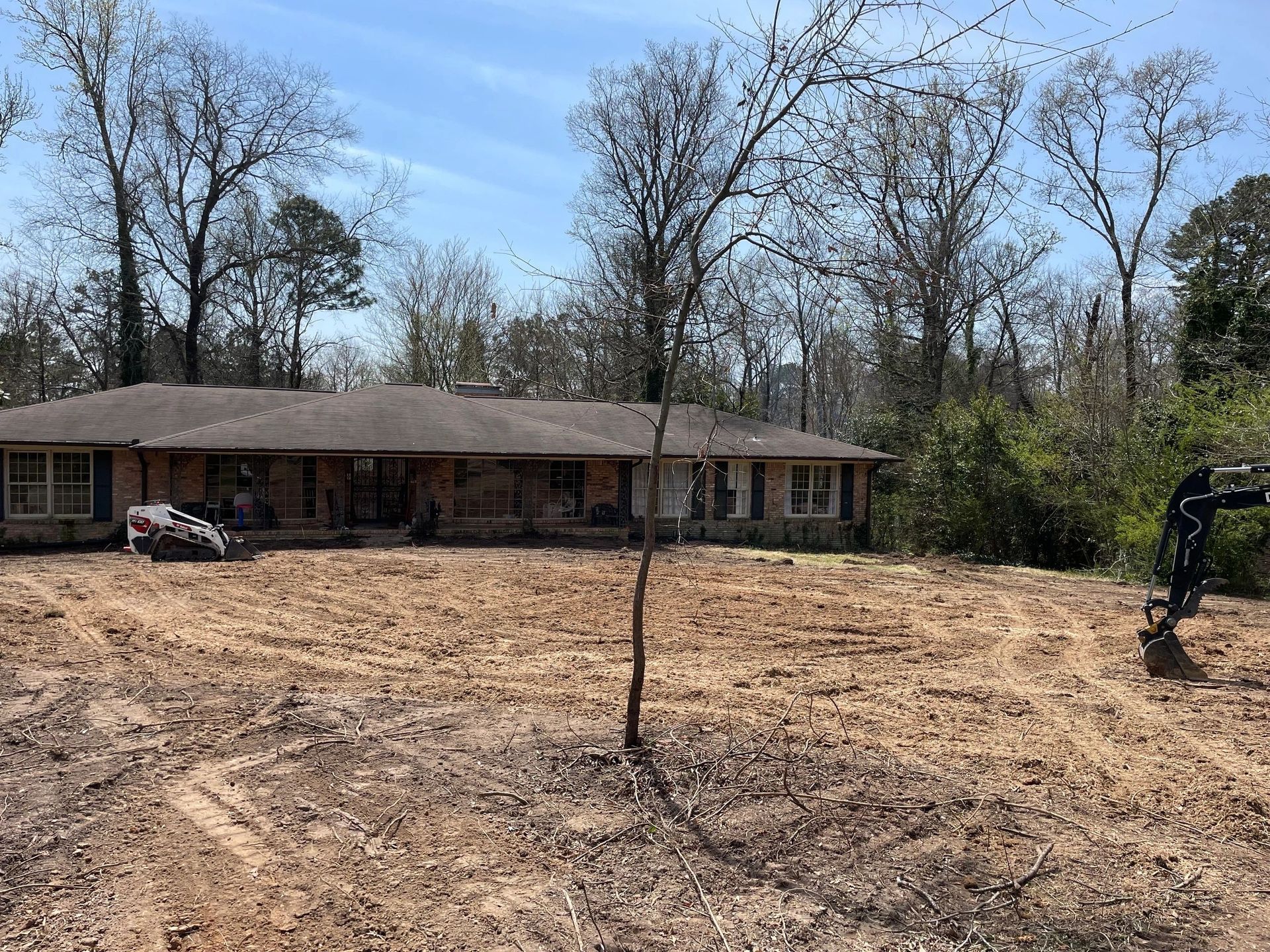 A low-slung house with a dark roof stands behind a freshly cleared lot of bare, dirt-covered ground under a clear sky.