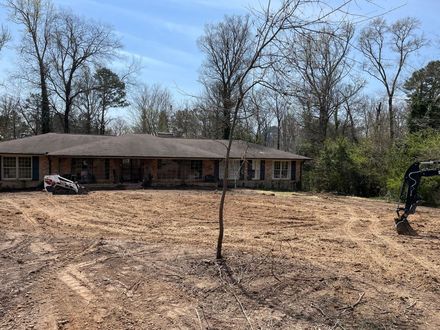 A house with a low-pitched roof sits behind a large area of cleared, brown dirt, with woods in the background.