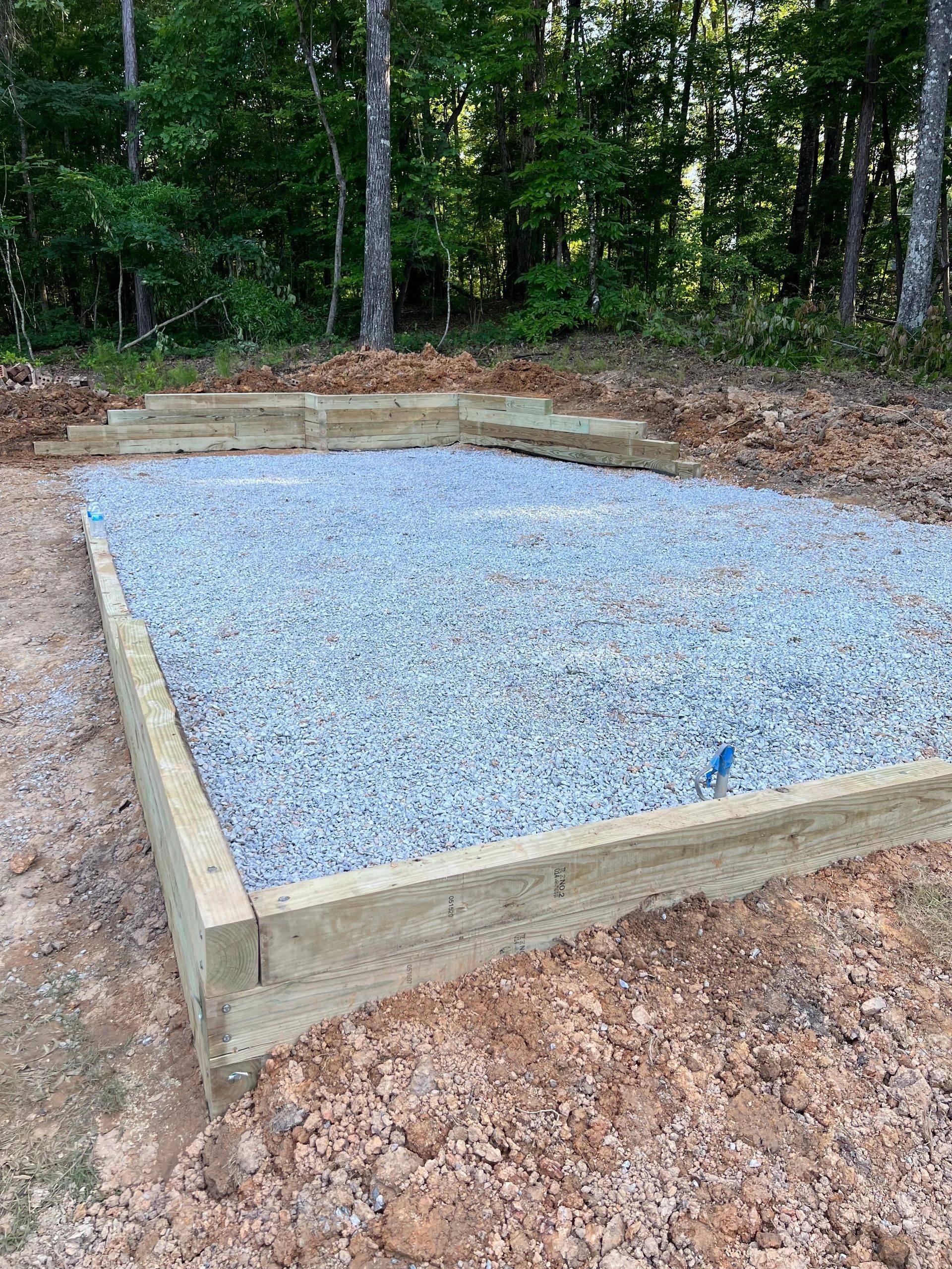 A leveled, rectangular gravel foundation framed with light-colored timber beams set in a wooded area.