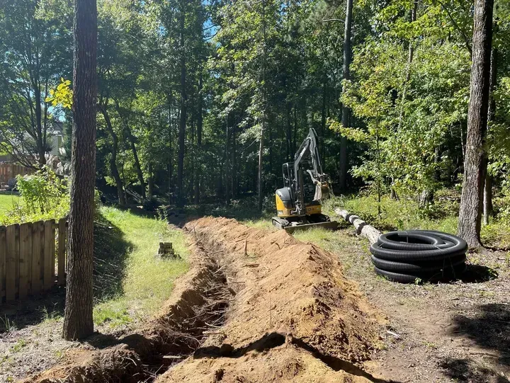 A yellow excavator sits in a wooded yard near a long, excavated trench and a coil of black corrugated drainage pipe.