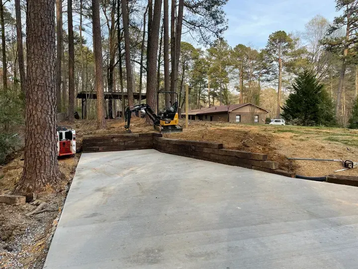 A newly poured concrete driveway bordered by a wooden retaining wall and pine trees in a wooded yard.