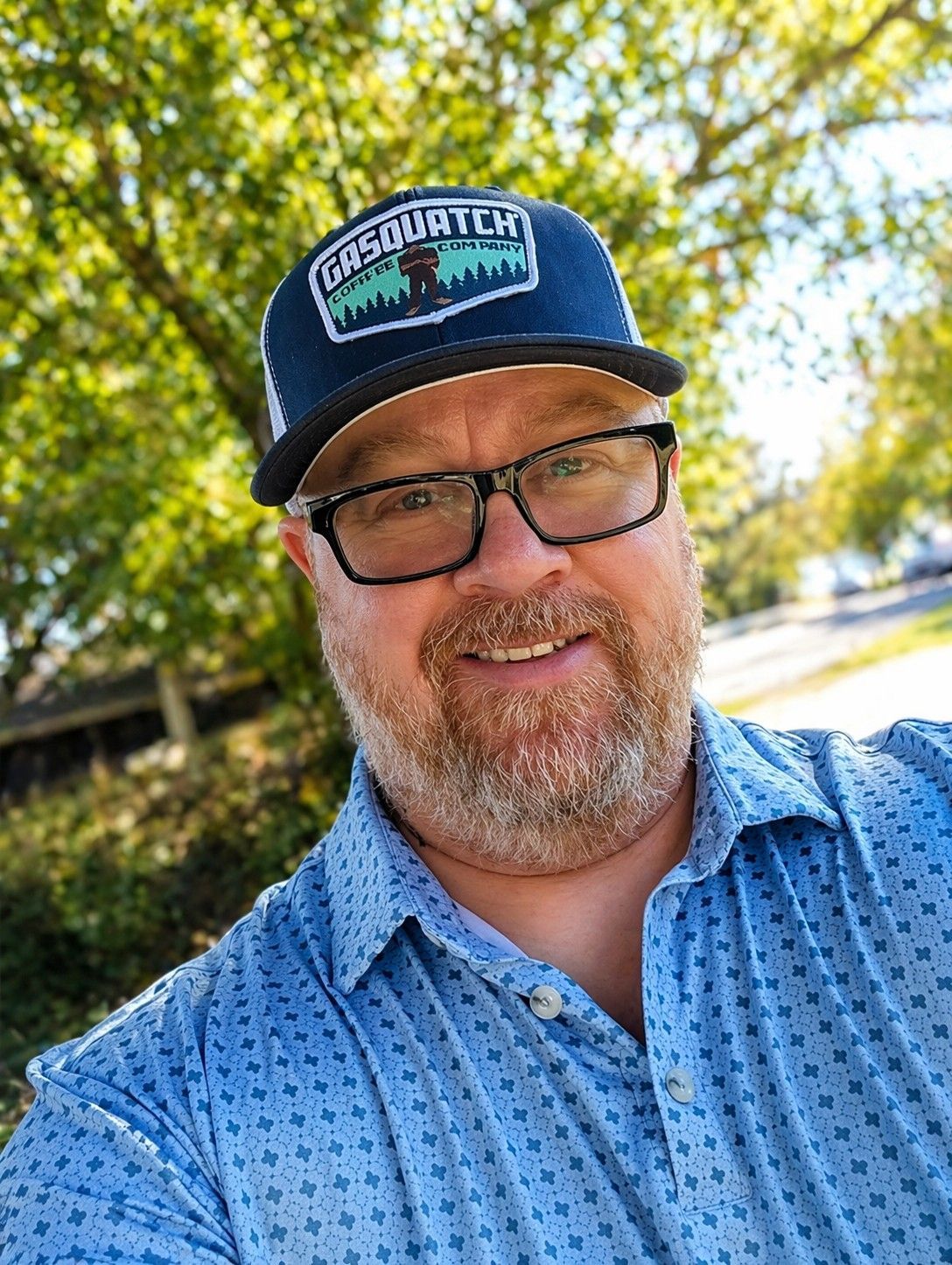 Smiling man in glasses and a navy cap outdoors in a blue patterned shirt