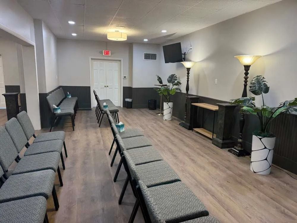 Empty waiting room with rows of gray chairs, white walls, potted plants, and a wall-mounted TV.