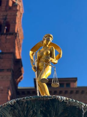 A golden statue of Lady Justice holding scales, positioned above a fountain with a brick tower in the background.
